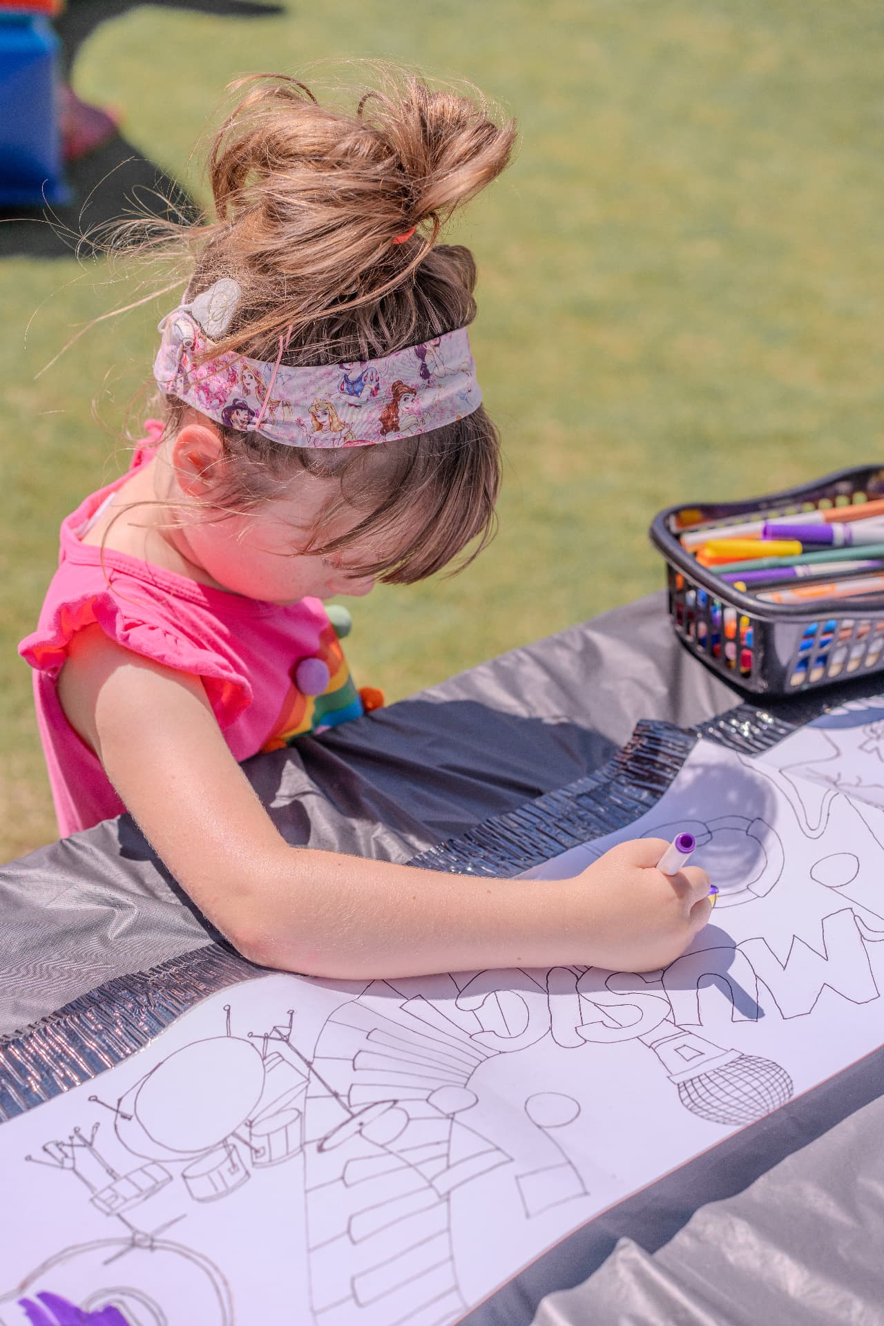 A young girl coloring a giant MUSIC mural with markers at an outdoor art table